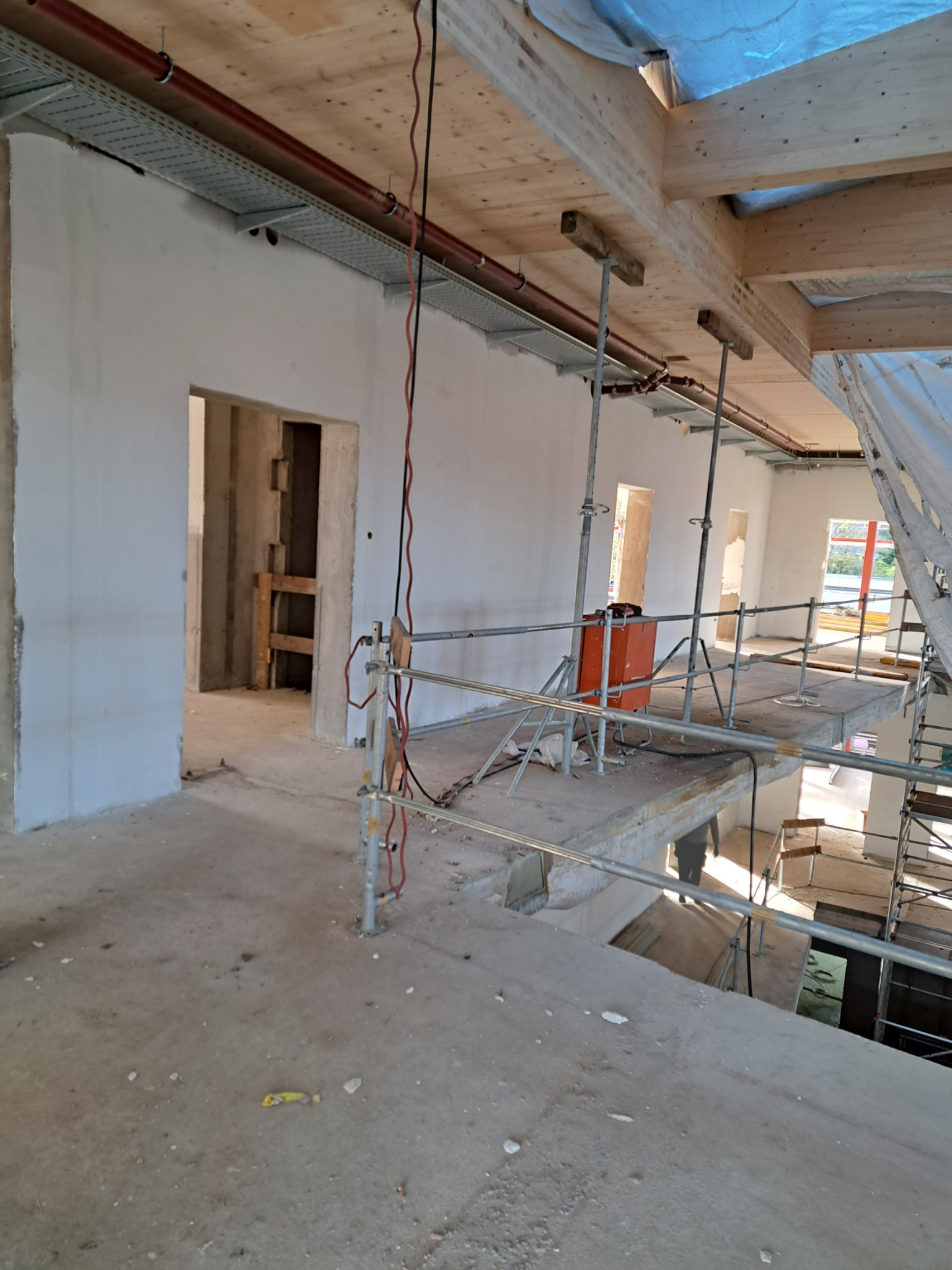 Wnętrze w trakcie prac wykończeniowych; widoczne surowe ściany, drewniana konstrukcja stropu, instalacje pod sufitem, rusztowania i otwarte przejścia między pomieszczeniami.