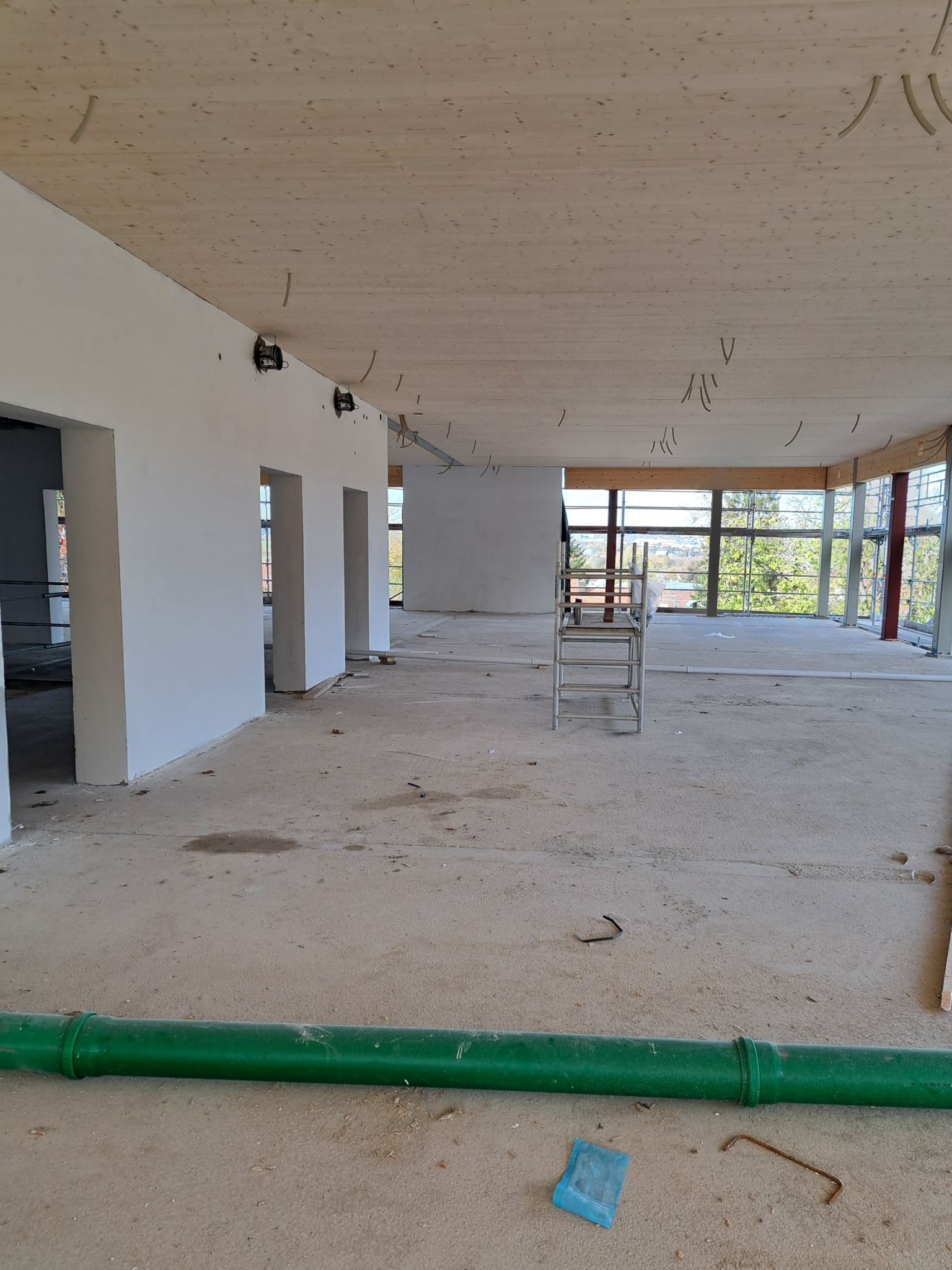 Surowe wnętrze w trakcie prac wykończeniowych, z drewnianym sufitem, białymi ścianami, zielonymi rurami na podłodze i aluminiową drabiną w tle okna.