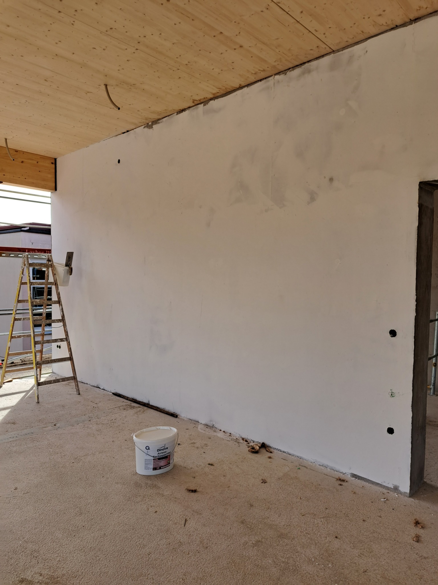 Ściana w trakcie szpachlowania, widoczny biały grunt, drabina z narzędziami i wiadro z masą szpachlową Dinova Airless Spachtel, drewniany sufit z widocznymi kablami.