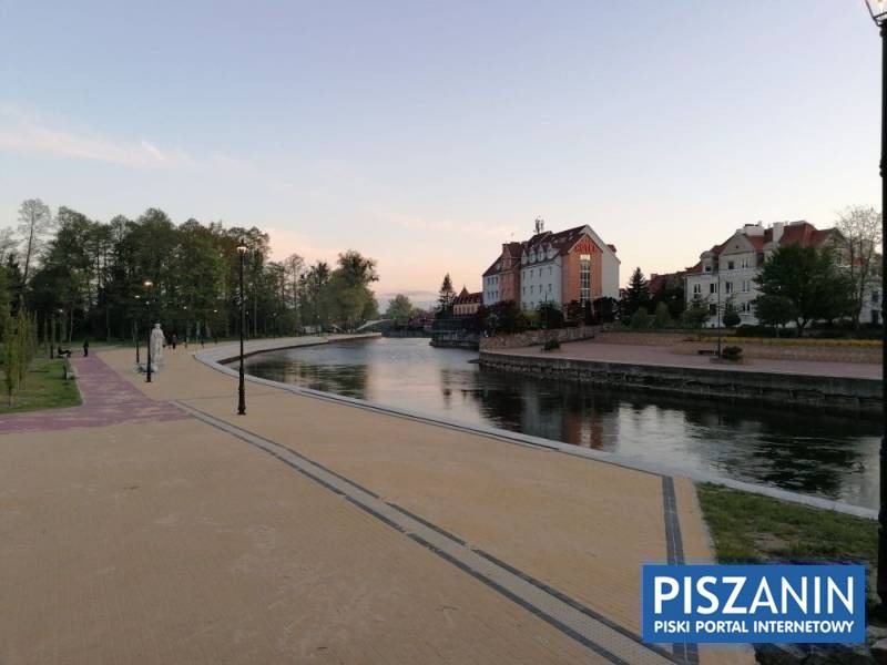 Nadbrzeżna promenada z widokiem na rzekę, budynki w tle i rzeźbę po lewej stronie, oświetlona latarniami, wczesnym wieczorem.