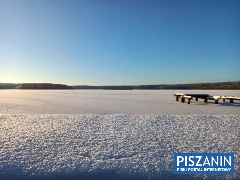 Zimowy krajobraz: Zamarznięte jezioro z drewnianym pomostem, pokryte śniegiem, las na horyzoncie, błękitne niebo.