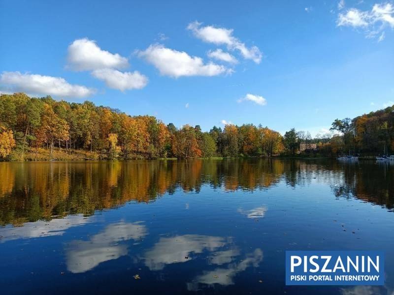 Jesienny krajobraz: jezioro z odbiciem nieba i chmur, otoczone lasem w jesiennych barwach, w tle widoczny budynek.