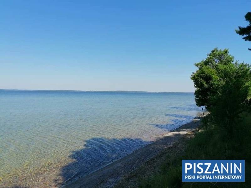 Spokojne wody jeziora z widocznym brzegiem, zieleń drzew po prawej stronie i logo 'PISZANIN PISKI PORTAL INTERNETOWY' w prawym dolnym rogu.