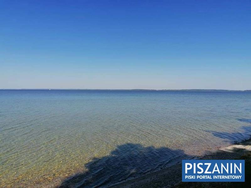 Szeroki kadr na spokojną taflę jeziora w słoneczny dzień, z widocznym dnem w strefie przybrzeżnej i logo 'PISZANIN' w prawym dolnym rogu.