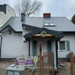 Dom jednorodzinny z nowym, szarym dachem z blachy falistej, widoczne okna dachowe i komin z cegły, dekoracyjny wieniec nad wejściem, skrzynka na listy i tabliczka z numerem 5 przy elewacji...