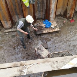 GGS-Projekt Laboratorium geotechniczne Sp. z o.o. - Pracownik w kasku pobiera próbki gruntu w wykopie zabezpieczonym stalowymi ściankami Larsena, na palecie leży niebieska folia i rękawice robocze.