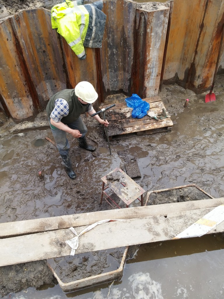 Pracownik w kasku pobiera próbki gruntu w wykopie zabezpieczonym stalowymi ściankami Larsena, na palecie leży niebieska folia i rękawice robocze.