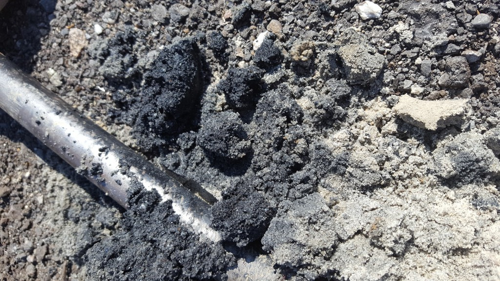Próbka gruntu składająca się z ciemnego, drobnoziarnistego materiału i jasnego piasku, naruszona metalowym prętem, sugerująca analizę laboratoryjną.