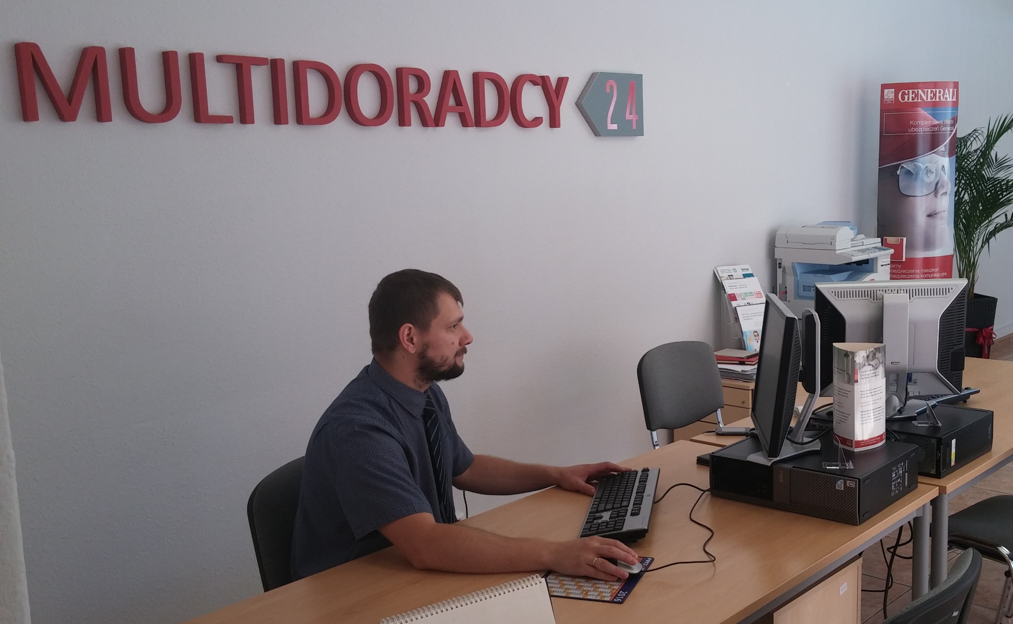Mężczyzna w biurze agencji ubezpieczeniowej Multidoradcy24, siedzący przy biurku z komputerem, w tle baner reklamowy Generali i materiały informacyjne.