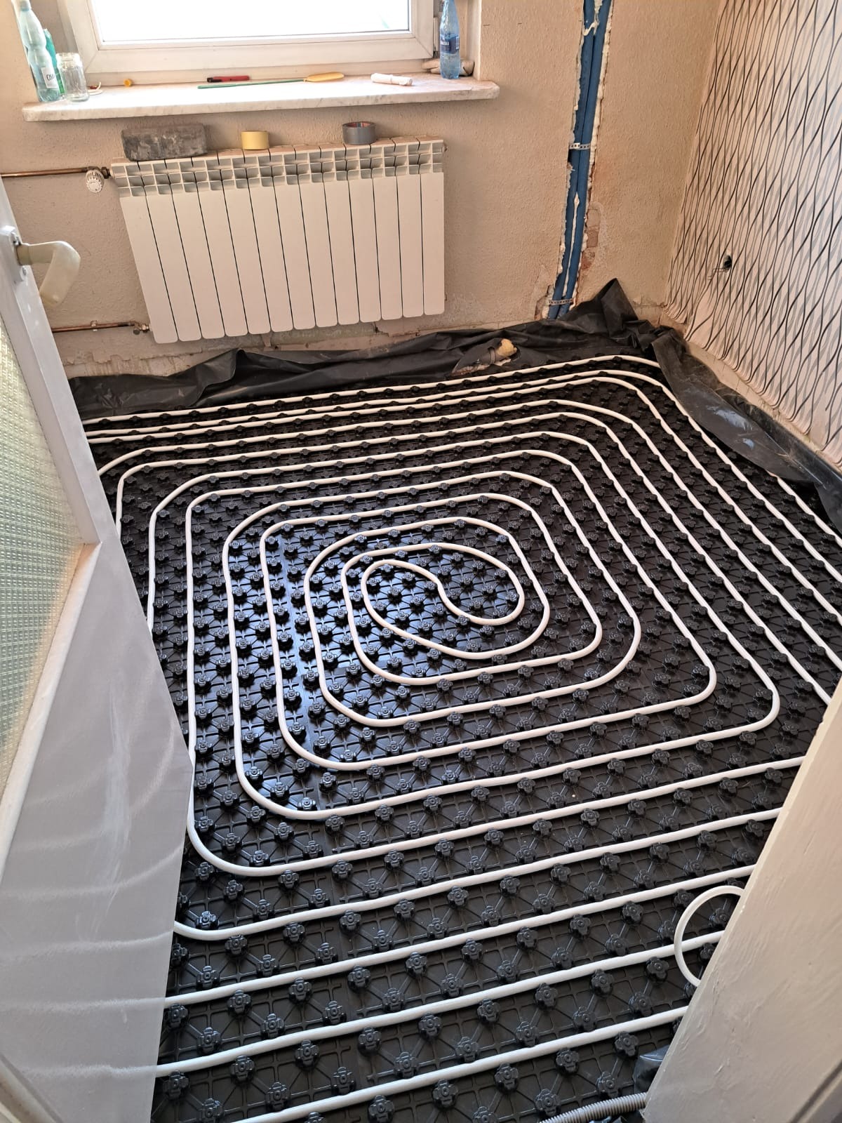 Białe rurki ogrzewania podłogowego ułożone spiralnie na czarnej macie izolacyjnej w trakcie montażu, widoczny grzejnik na ścianie i fragment okna.