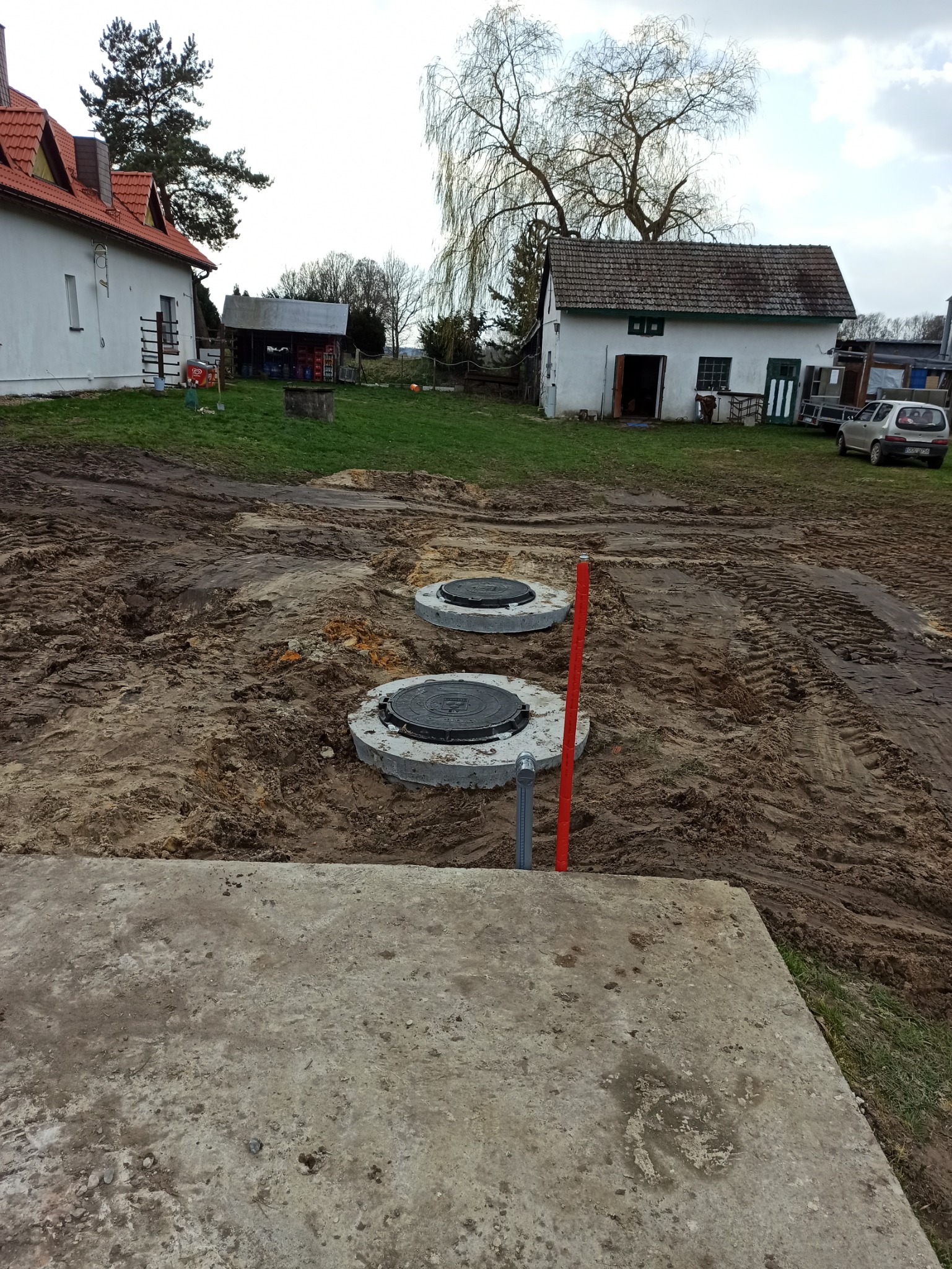 Dwa betonowe kręgi studni kanalizacyjnej z pokrywami, częściowo zakopane w ziemi na posesji w Chudobie. W tle budynki gospodarcze i trawnik.