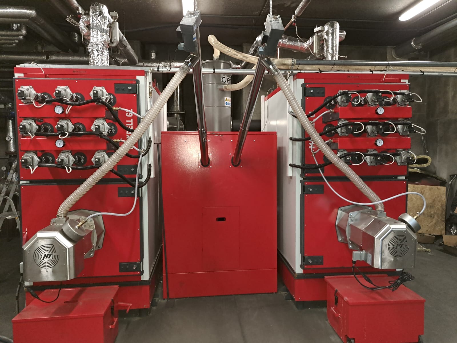 Kompleksowa kotłownia z dwoma czerwonymi kotłami i srebrnym zbiornikiem wyrównawczym, widoczne rury i manometry, całość w pomieszczeniu technicznym.