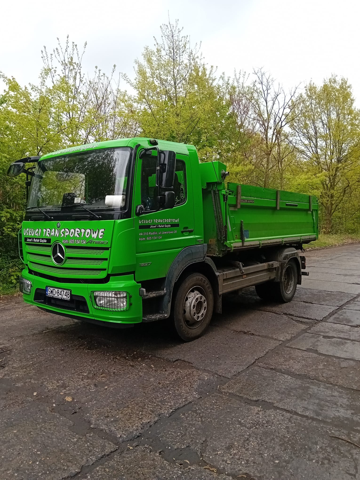 Jasnozielona wywrotka Mercedes z napisem 'Usługi Transportowe' zaparkowana na betonowej nawierzchni, w tle drzewa.