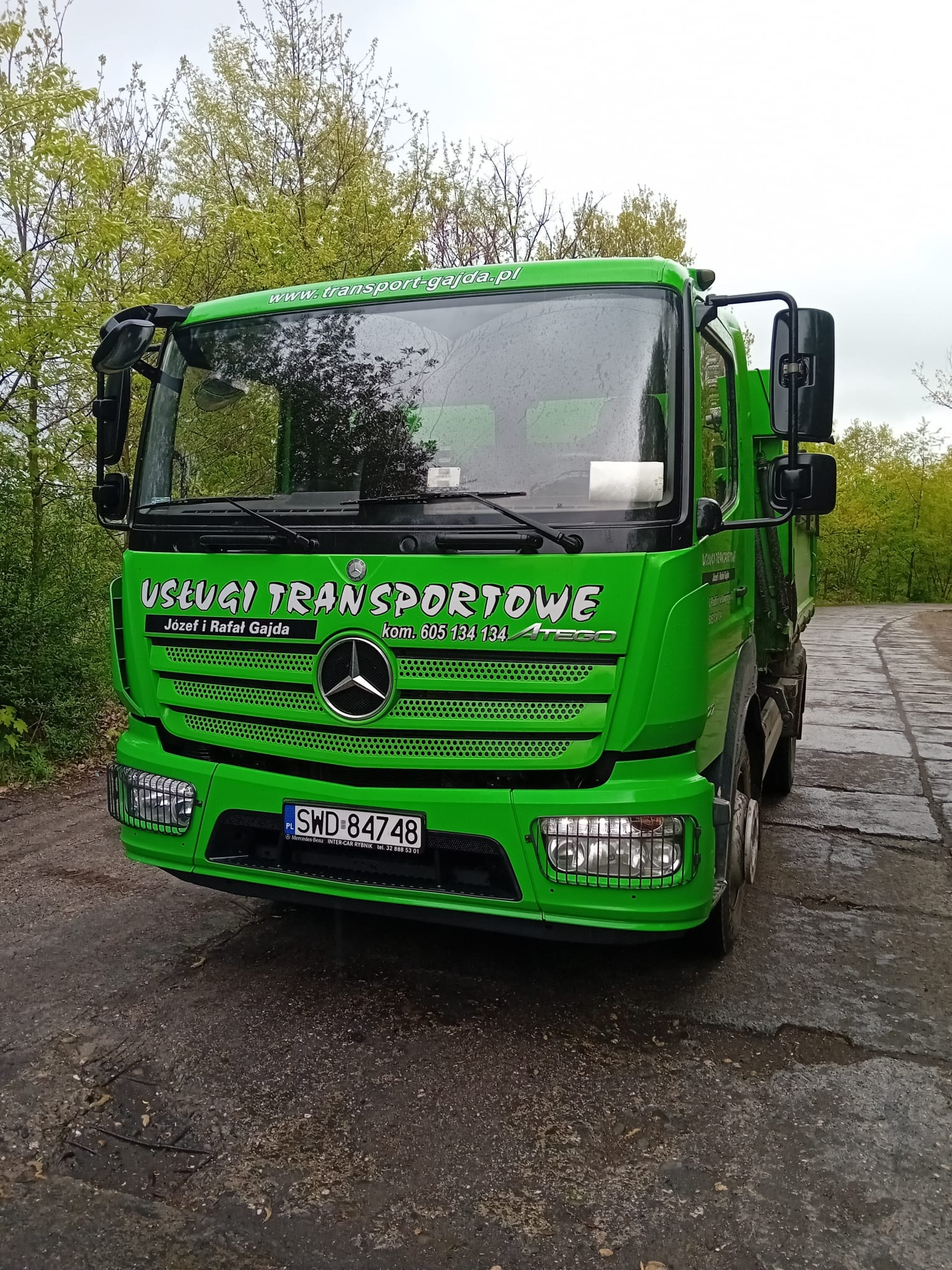 Zielony samochód ciężarowy marki Mercedes-Benz Atego z napisem 'Usługi Transportowe Józef i Rafał Gajda' na tle drzew i szarej drogi.