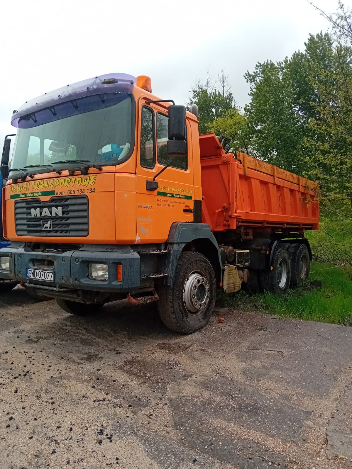 Pomarańczowa wywrotka marki MAN z widocznymi zabrudzeniami na karoserii i oponach, zaparkowana na poboczu drogi z zielonymi drzewami w tle.