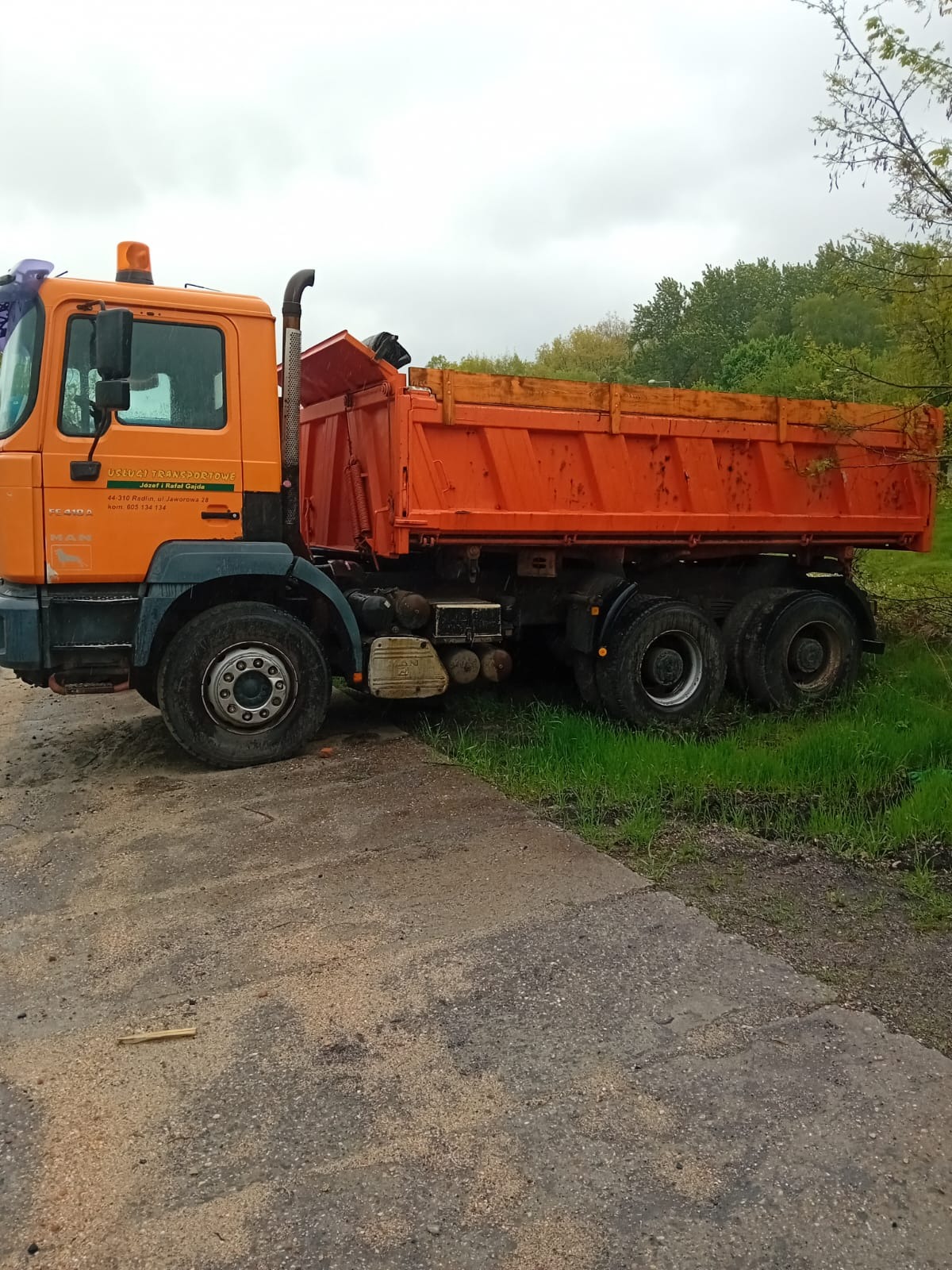 Pomarańczowa wywrotka MAN z widocznymi napisami 'USŁUGI TRANSPORTOWE Józef i Rafał Gajda' na kabinie, stojąca na betonowej nawierzchni z trawą w tle.