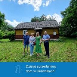 Blue Home Biuro Nieruchomości Chełm - Grupa czterech osób stoi przed żółto-brązowym, parterowym domem z ciemnym dachem na zarośniętej trawie, pozując do zdjęcia w słoneczny dzień.