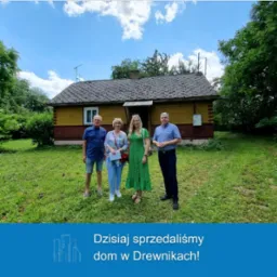 Grupa czterech osób stoi przed żółto-brązowym, parterowym domem z ciemnym dachem na zarośniętej trawie, pozując do zdjęcia w słoneczny dzień.