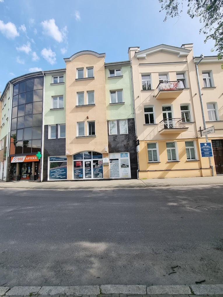 Fasada budynku z biurami i apteką przy ulicy Narutowicza w Chełmie, widoczne tablice informacyjne i okna wystawowe.