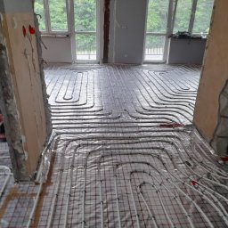 Lidia Turczyn-Bąk - Usługi Hydrauliczno - Budowlane - Instalacja ogrzewania podłogowego w trakcie montażu, widoczne rury grzewcze ułożone na folii izolacyjnej z siatką, surowy stan wykończenia wnętrza.