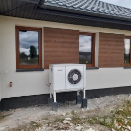 Lidia Turczyn-Bąk - Usługi Hydrauliczno - Budowlane - Biała pompa ciepła zamontowana na zewnątrz domu z brązowymi oknami i drewnianą elewacją, widok z poziomu gruntu.