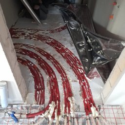 Lidia Turczyn-Bąk - Usługi Hydrauliczno - Budowlane - Montaż instalacji ogrzewania podłogowego: czerwone rury grzewcze mocowane do folii aluminiowej z użyciem pianki montażowej, widoczni instalatorzy w tle.