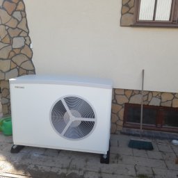 Lidia Turczyn-Bąk - Usługi Hydrauliczno - Budowlane - Biała pompa ciepła Stiebel Eltron zamontowana przy ścianie budynku z kamienną okładziną, widoczne zielone konewki i wiadro obok urządzenia.