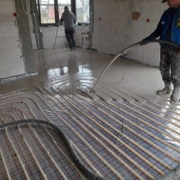 Lidia Turczyn-Bąk - Usługi Hydrauliczno - Budowlane - Wylewka anhydrytowa rozprowadzana na instalacji ogrzewania podłogowego w surowym wnętrzu, widoczny pracownik z wężem i drugi plan z oknem.