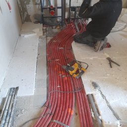Lidia Turczyn-Bąk - Usługi Hydrauliczno - Budowlane - Instalacja rur grzewczych w bruzdach podłogi, widoczny pracownik w trakcie montażu, narzędzia i elementy instalacji sanitarnej Grohe.
