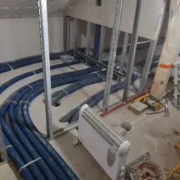 Niebieskie rury instalacji ogrzewania podłogowego ułożone w surowym stanie, tymczasowy grzejnik elektryczny w pomieszczeniu ze stelażem pod zabudowę, narzędzia w tle.