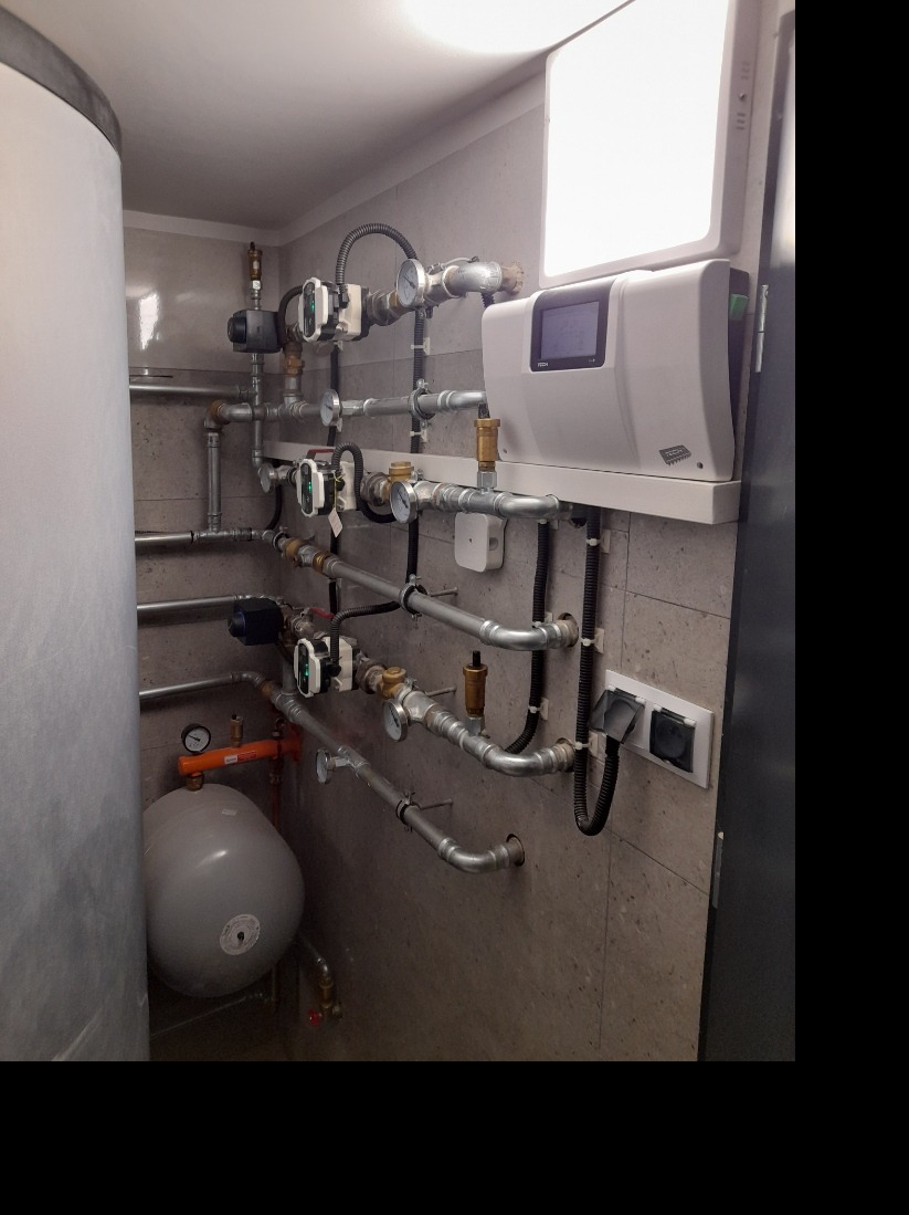 Węzeł cieplny w kotłowni: zbiornik akumulacyjny, liczne rury z zaworami i manometrami, elektroniczny regulator temperatury oraz biała lampa oświetlająca instalację.