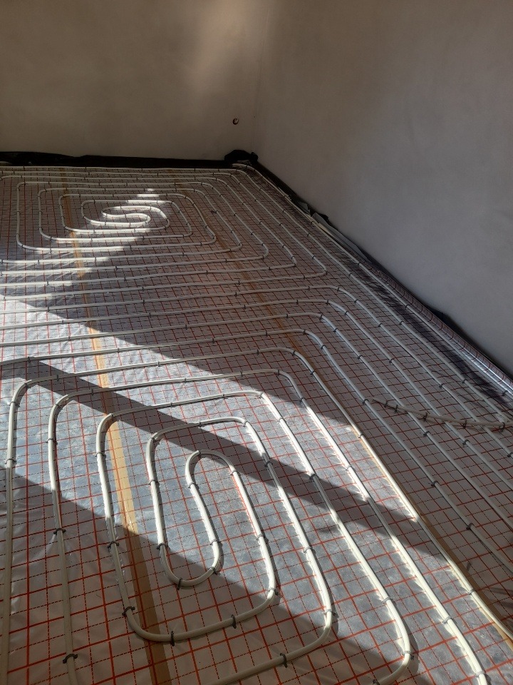 Białe rury ogrzewania podłogowego ułożone spiralnie na czerwonej siatce izolacyjnej w rogu pokoju, widoczne czarne taśmy izolacyjne przy ścianie.