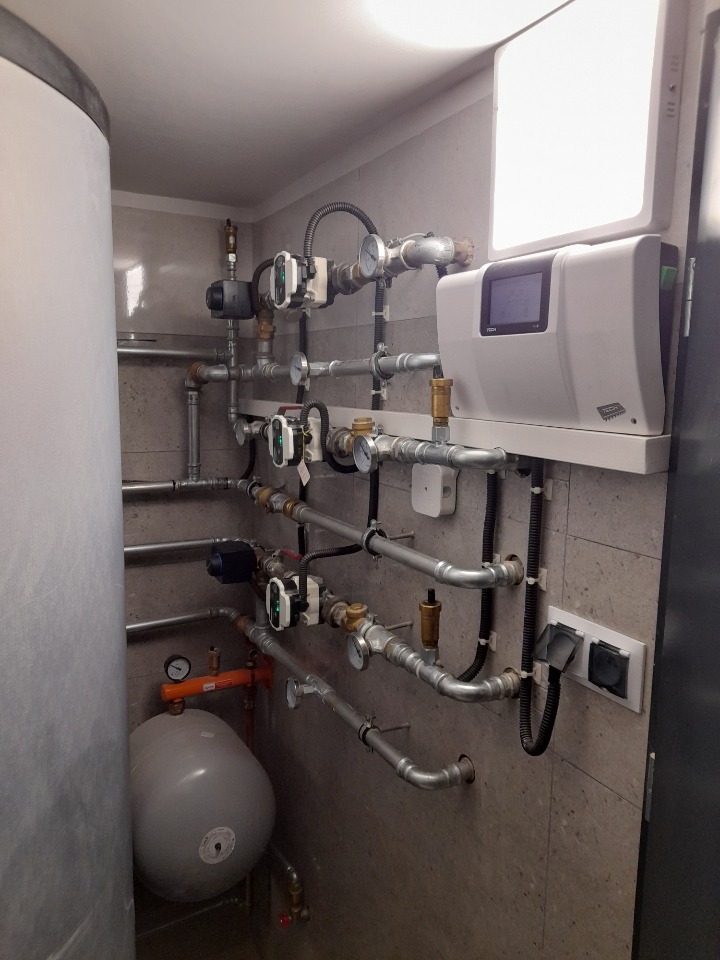 Złożona instalacja rur, zaworów, manometrów i pompy obiegowej w kotłowni, widoczny zbiornik akumulacyjny i sterownik systemu na ścianie z szarych płytek.