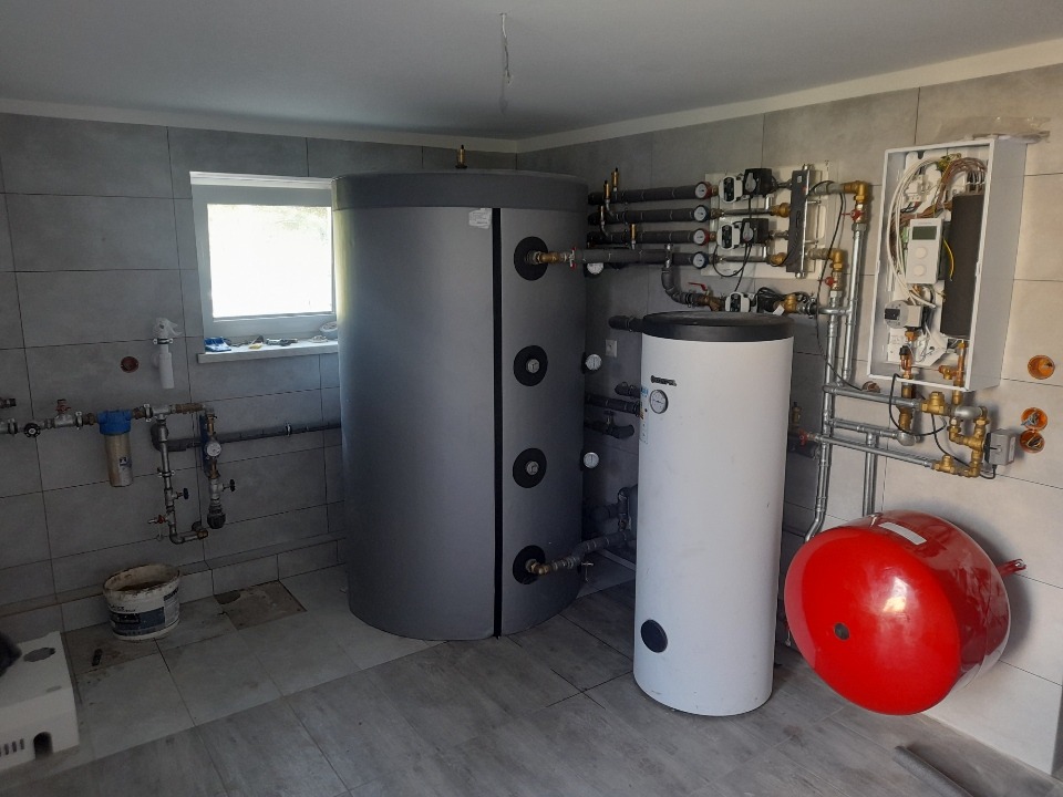 Zainstalowany system ogrzewania z dwoma zbiornikami akumulacyjnymi: szarym i białym oraz czerwonym naczyniem przeponowym w kotłowni w Opolu Lubelskim, widoczne liczne rury i zawory.