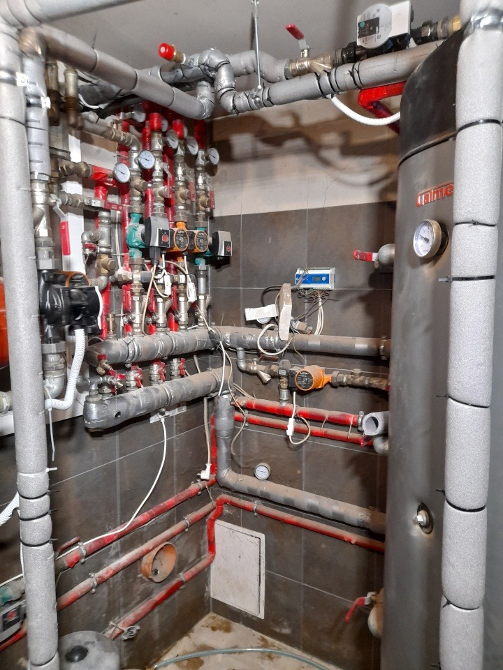 Złożona instalacja rur grzewczych i wodnych z licznymi zaworami, manometrami i pompami, częściowo zaizolowana, z widocznym zasobnikiem ciepłej wody w kotłowni wyłożonej ciemnymi płytkami.