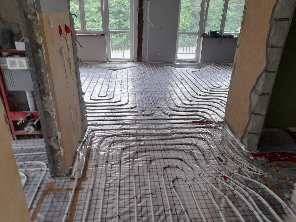 Instalacja ogrzewania podłogowego w trakcie montażu, widoczne rury grzewcze ułożone na folii izolacyjnej z siatką, surowy stan wykończenia wnętrza.