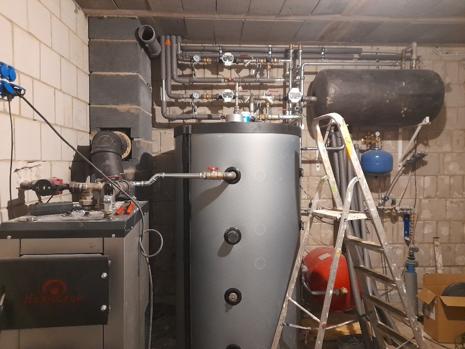 Piwnica z kotłem na paliwo stałe, zasobnikiem ciepłej wody użytkowej i rozbudowaną instalacją hydrauliczną, widoczna drabina i narzędzia.