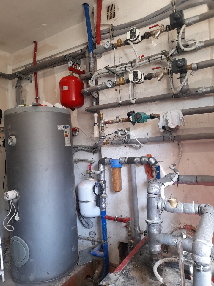 Skomplikowana instalacja grzewcza z licznymi rurami, zaworami, pompami obiegowymi, zbiornikiem wyrównawczym i filtrem wody, widoczna w pomieszczeniu technicznym.