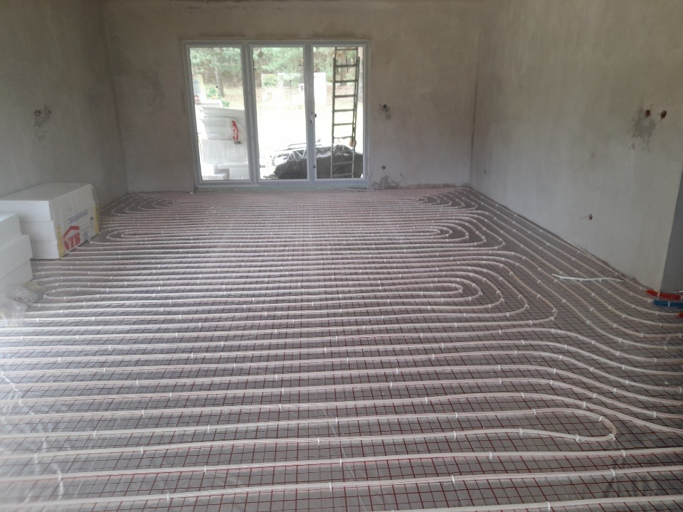 Niedokończona instalacja ogrzewania podłogowego w surowym pomieszczeniu, rury ułożone na siatce, widoczne materiały izolacyjne przy oknie.