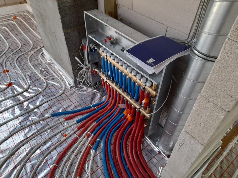Rozdzielacz ogrzewania podłogowego z podłączonymi rurami w kolorach czerwonym i niebieskim, widoczna instalacja na folii izolacyjnej oraz notatnik na obudowie rozdzielacza.