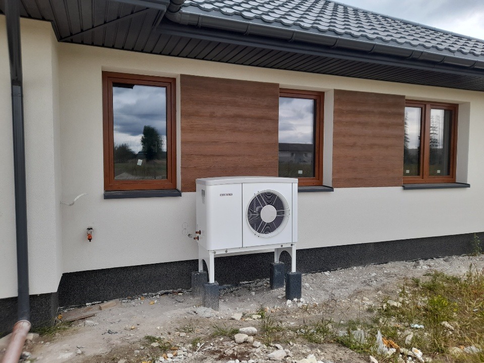Biała pompa ciepła powietrze-woda, zamontowana na zewnątrz domu jednorodzinnego z brązowymi oknami i elewacją w odcieniach bieli i szarości, widoczna instalacja przyłączeniowa.
