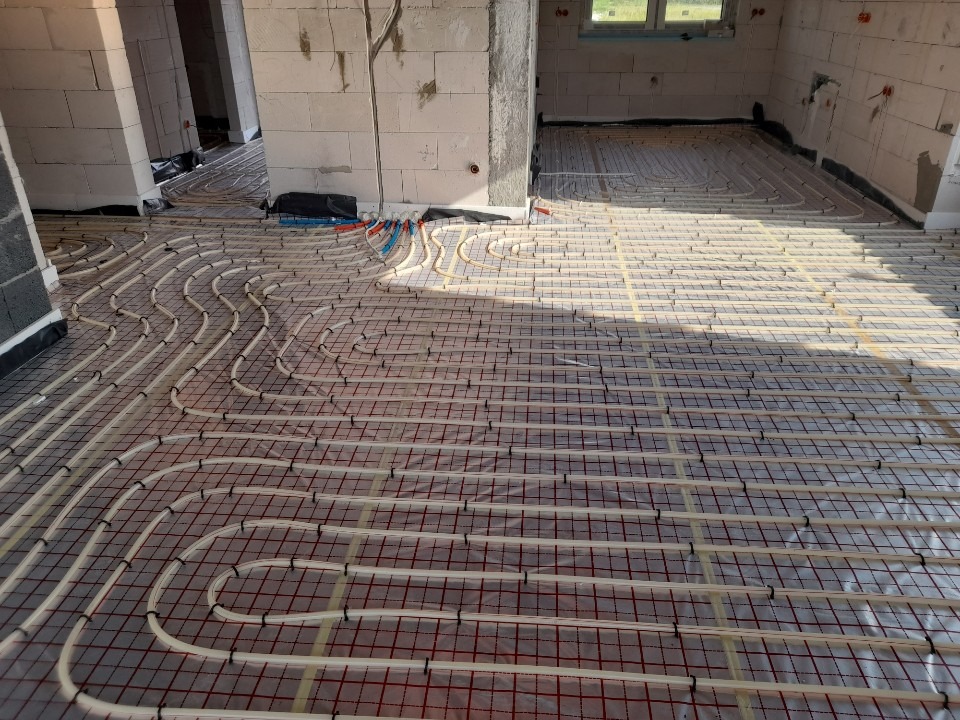 Rozłożone rury ogrzewania podłogowego na folii z rastrem w surowym wnętrzu domu w trakcie budowy, widoczne białe bloczki ścian.