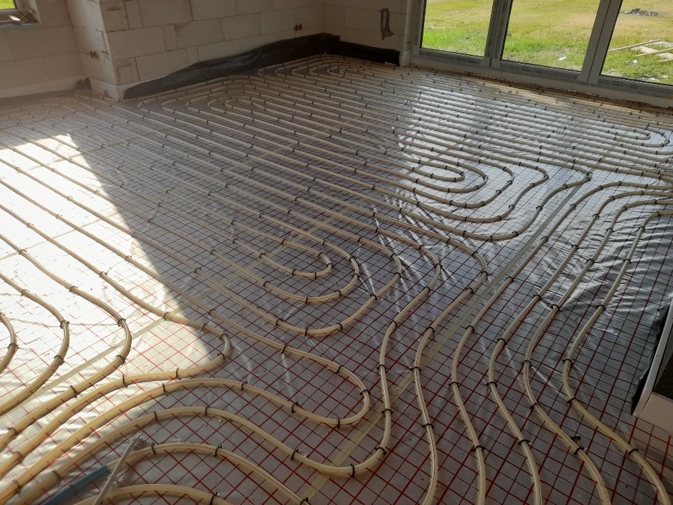 Instalacja ogrzewania podłogowego w surowym stanie, rury grzewcze ułożone na siatce, widoczne okno i fragment ściany z pustaków.