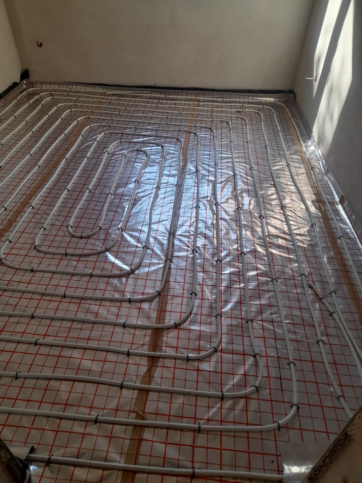 Białe rury grzewcze ułożone w pętle na folii z czerwoną kratką, przygotowane do zalania wylewką w jasnym pomieszczeniu.