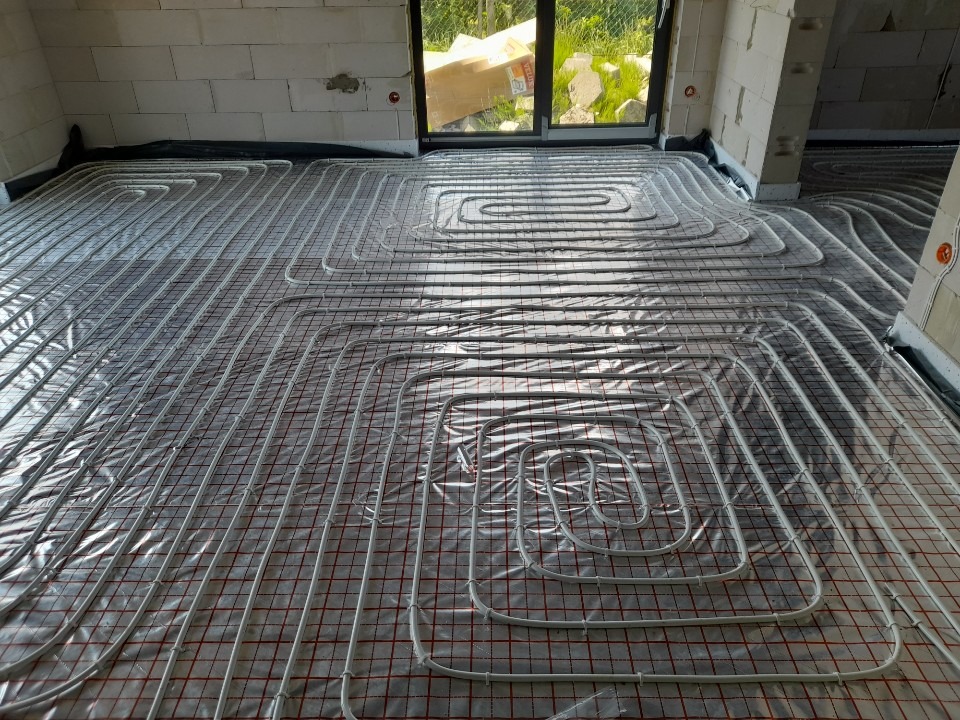 Białe rury ogrzewania podłogowego ułożone na folii z siatką w surowym wnętrzu budynku w trakcie budowy, widok z góry.