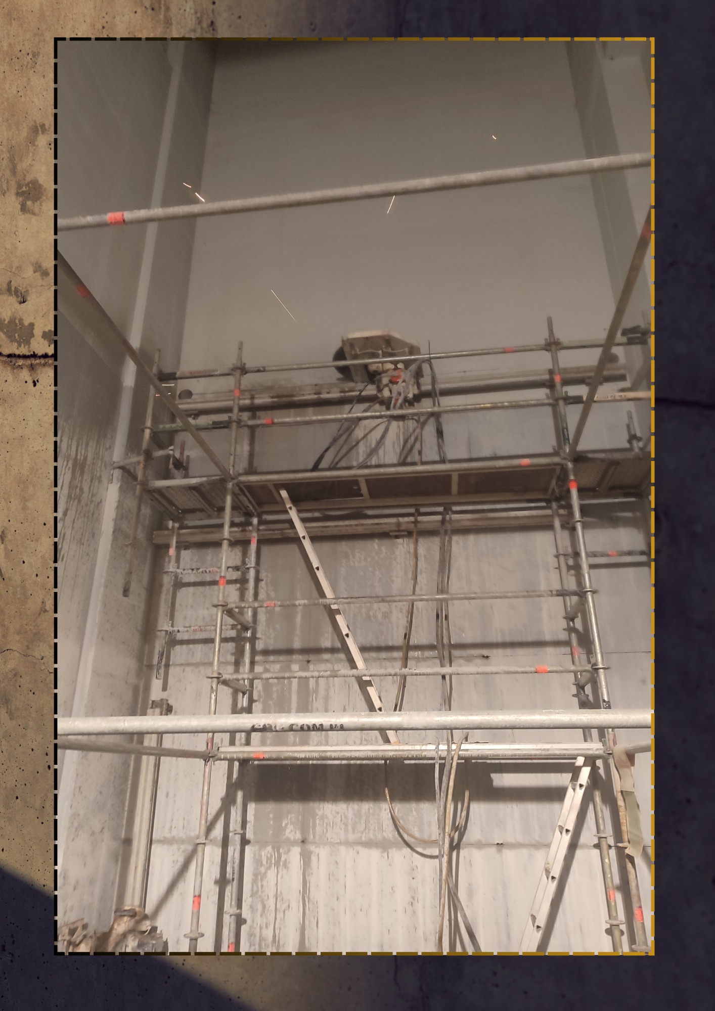 Wnętrze budynku w trakcie prac wyburzeniowych, widoczne rusztowanie i urządzenie do cięcia betonu na ścianie, drobne iskry podczas cięcia.