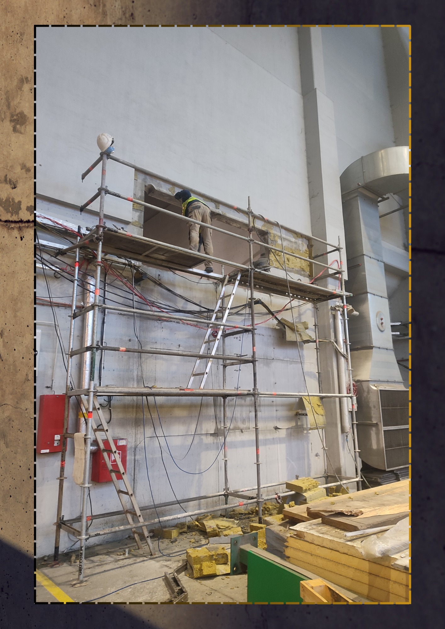 Pracownik w hełmie i kamizelce na rusztowaniu, wykuwający otwór w ścianie przemysłowego budynku, z widocznymi kablami, instalacjami i gruzem na podłodze.