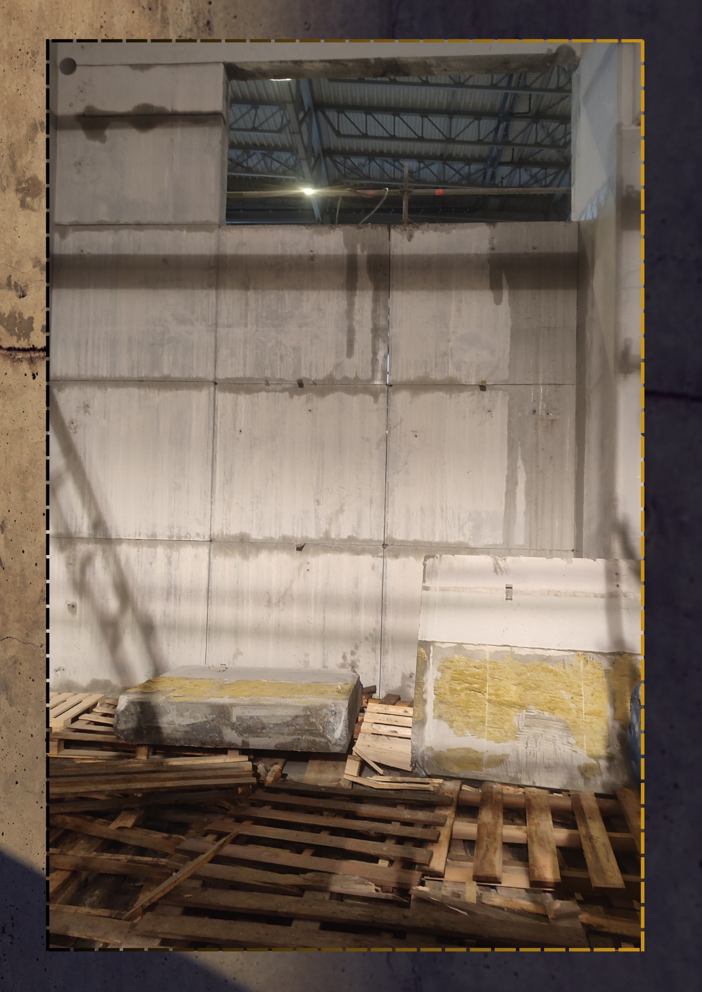 Wznoszenie betonowej ściany z prefabrykatów w hali magazynowej, widoczne palety i fragment izolacji.