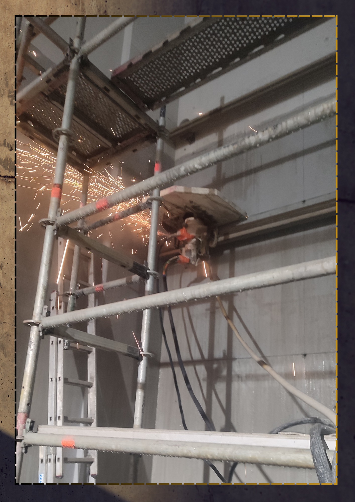 Cięcie betonu piłą diamentową zamontowaną na szynie, z iskrami, na tle szarej ściany, widoczne rusztowanie.