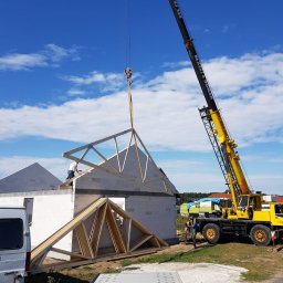 HONEST INVEST SP.ZO.O - Żółty dźwig unosi drewnianą konstrukcję dachu nad budynkiem z białych bloczków, pracownik na ścianie, błękitne niebo w tle.